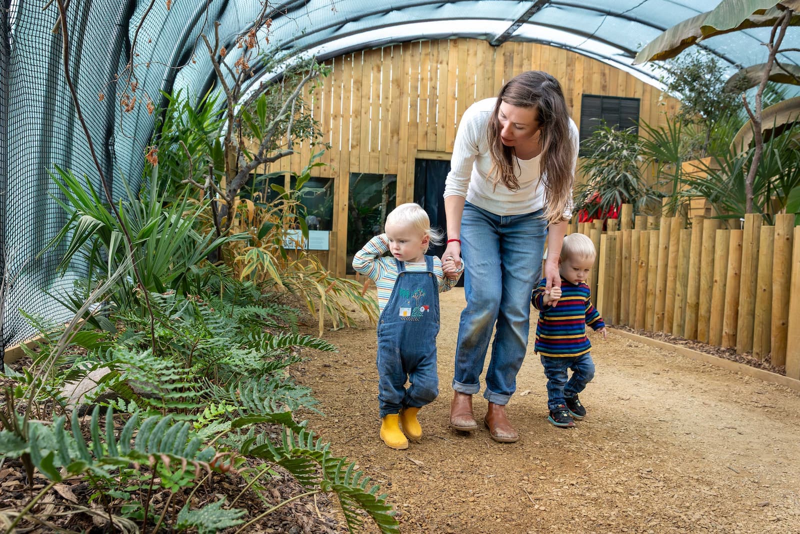 A woman walks with two toddlers on a path surrounded by plants in a greenhouse. The toddlers wear overalls and colorful shirts.