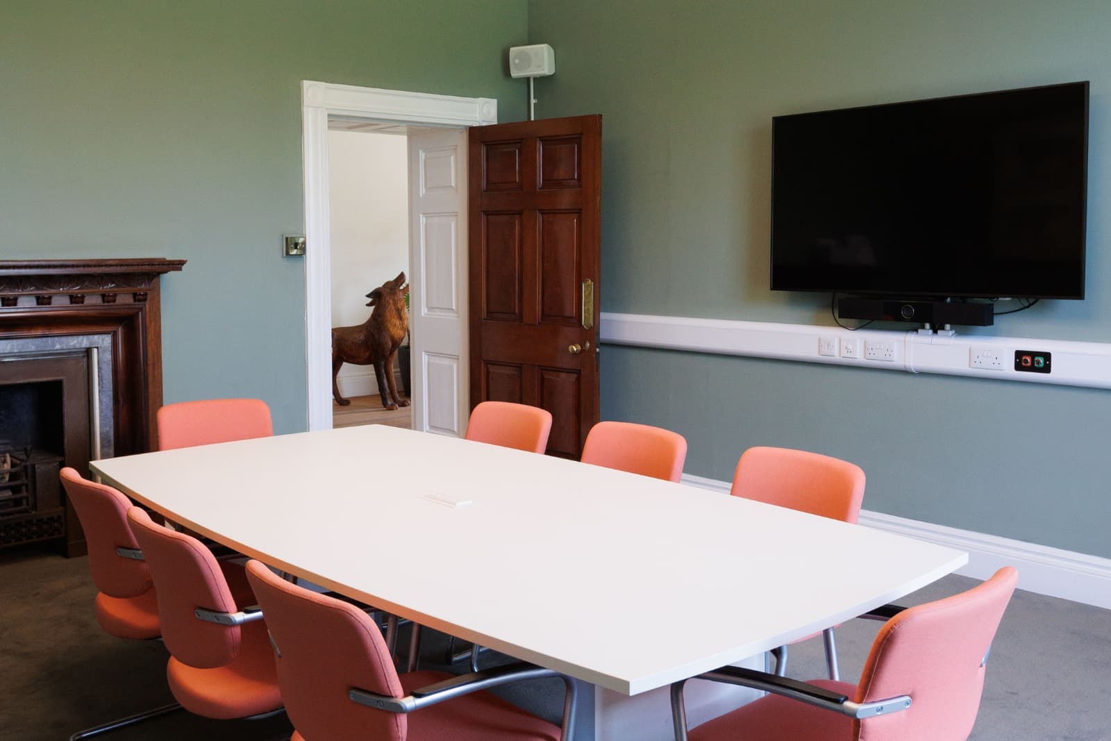 Conference room with white table and coral chairs, wall-mounted TV and fireplace; open wooden door reveals a life-size fox statue in the next room.