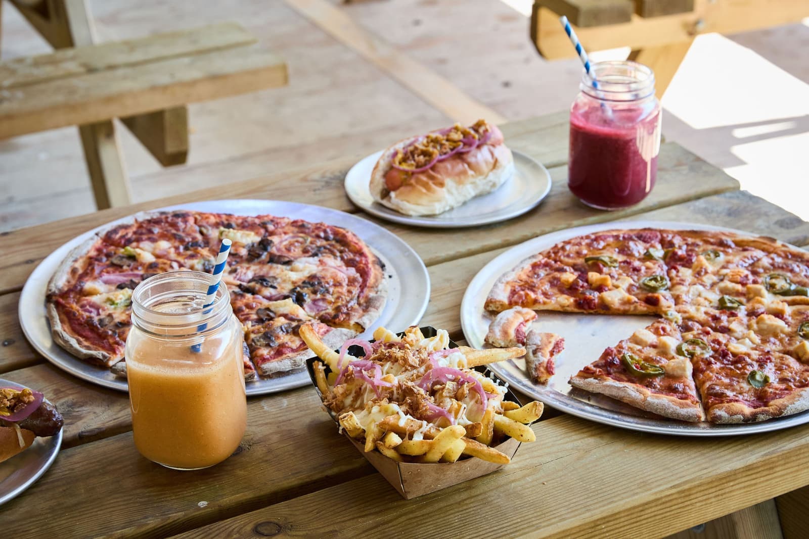 Pizzas, a hot dog, loaded fries, and smoothies on a wooden picnic table outdoors.